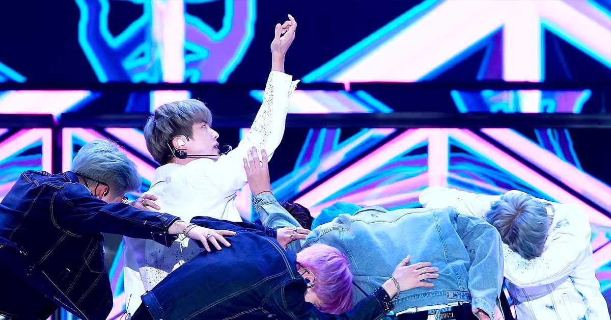 Nastop K-pop skupine BTS. Pet fantov na odru v plesni formaciji, kjer štirje klečijo in gledajo v tla, medtem ko eden izmed njih dviguje roko v zrak in gleda gor.