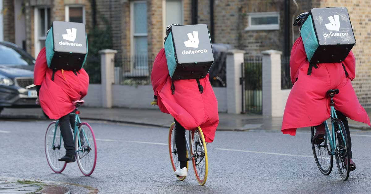 Trije dostavljalci v rdečih dežnih plaščih na kolesih, ki imajo na ramenih Deliveroo torbe.