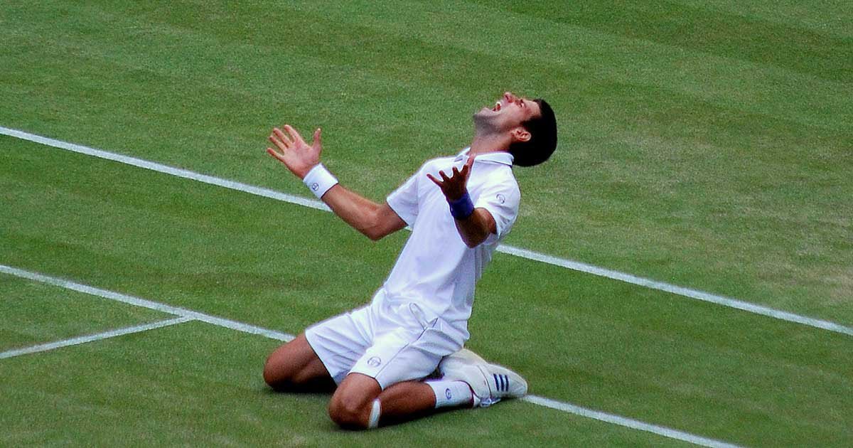 Novak Đoković kleči na teniškem igrišču in vpije v nebo.