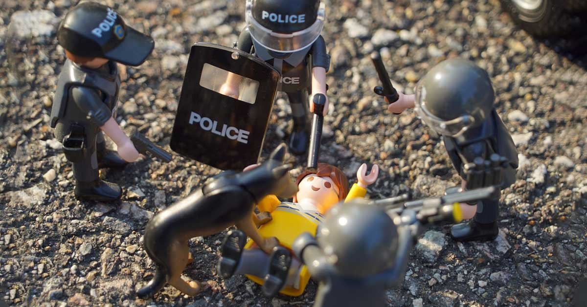 Simulacija policijskega nasilja z Lego figuricami. Štirje polno opremljeni policaji in policijski pes napadajo osebo, ki je na tleh.