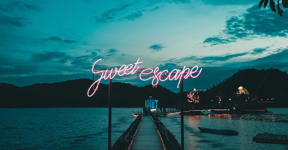 Morska idila ob mraku. Pot na vodi, ki vodi do objekta, nad vhodom poti je neonski napis Sweet escape.
