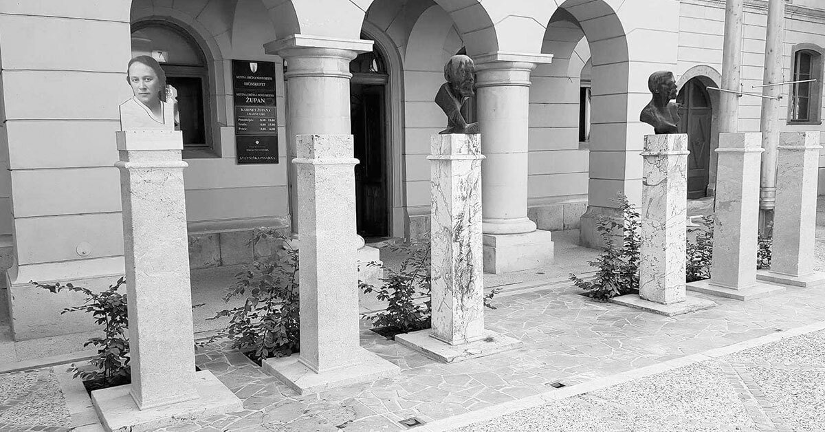 Na fotografiji je špalir kipov na glavnem novomeškem trgu, med katerimi so štiri prazna stojala. Na prvem je provizorično postavljena glava Ilke Vašte.