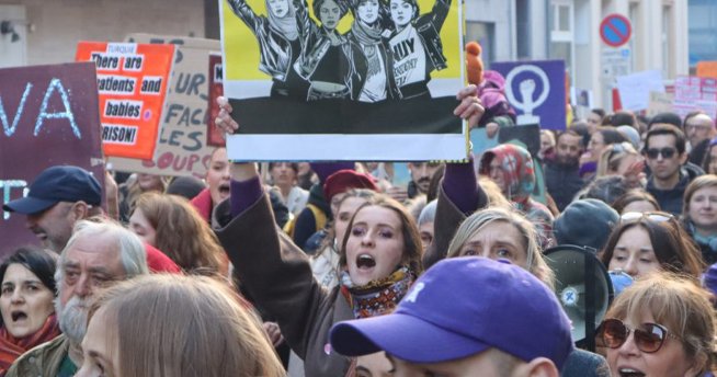 Fotografija iz protesta za pravico do splava, na kateri je veliko ljudi, v sredini pa ženska, ki drži plakat.