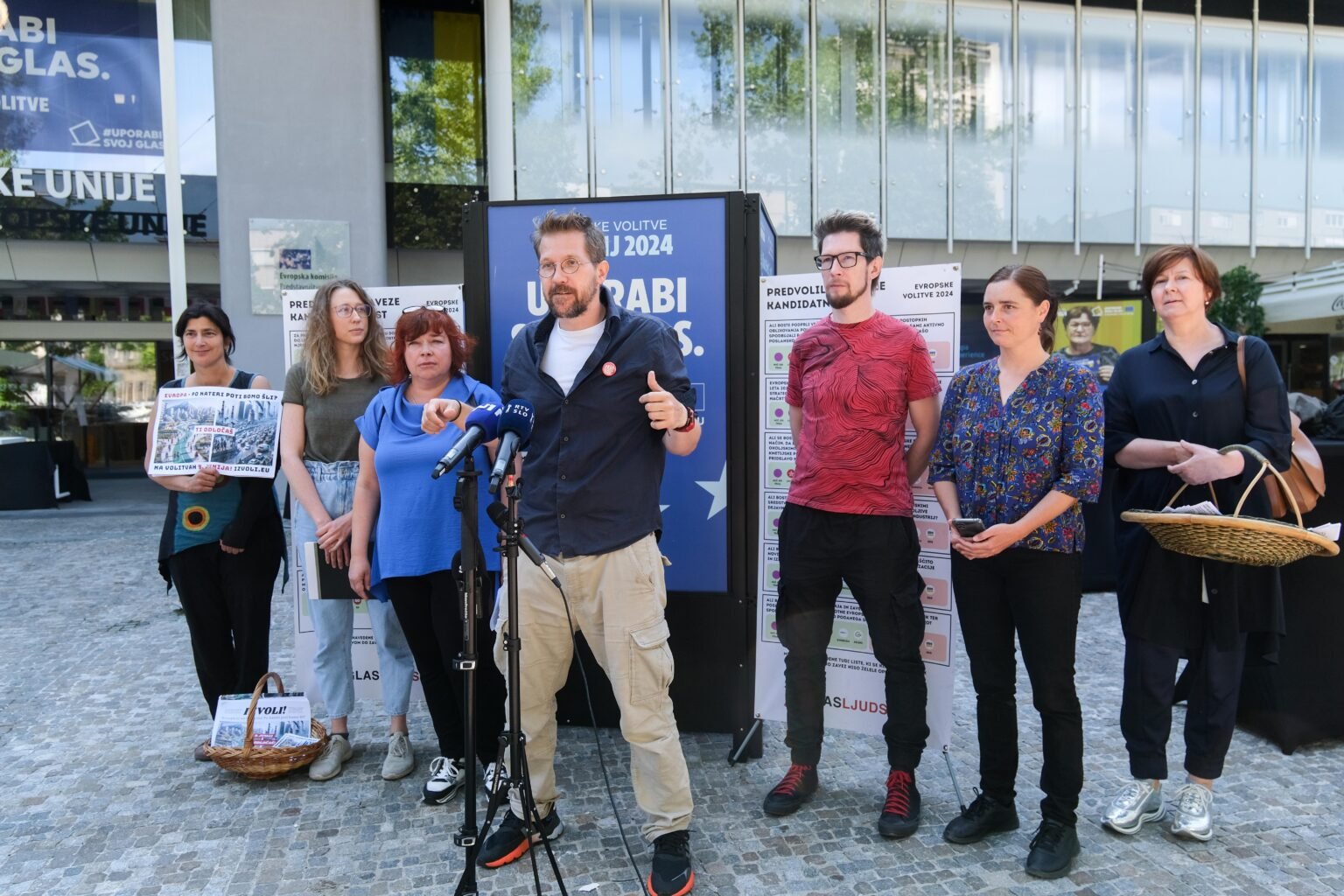 Novinarska konferenca Glas ljudstva. Vidimo 7 članov ekipe, 1 oseba je pred mikrofonom.