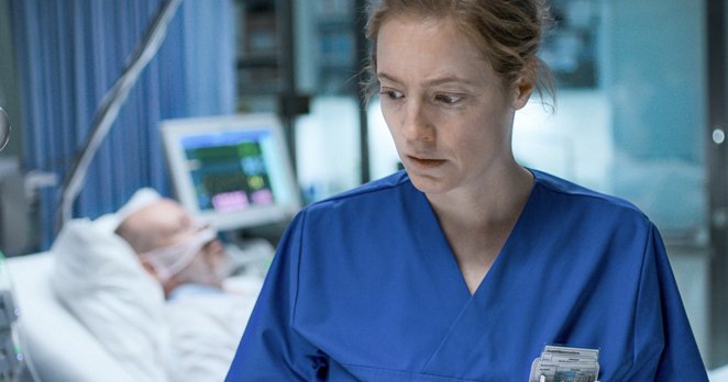 Zdravstvena delavka v modri uniformi stoji ob bolniški postelji v bolniški sobi; v ozadju leži pacient, priključen na medicinske naprave, na monitorju so vidni življenjski znaki.