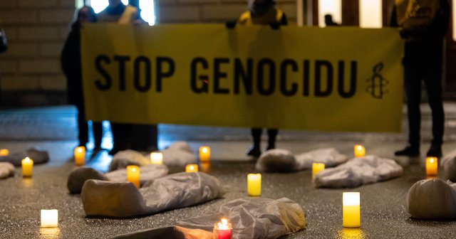 V ozadju transparent, na katerem piše STOP GENOCIDU, v ospredju na tleh sveče in vreče, ki simbolizirajo trupla.