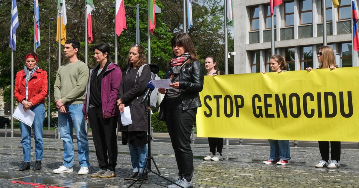 Fotografija predstavnic in predstavnika organizacij, ki so na novinarski konferenci pred DZ predstavile peticijo, v ozadju rumen transparent z napisom Stop genocidu