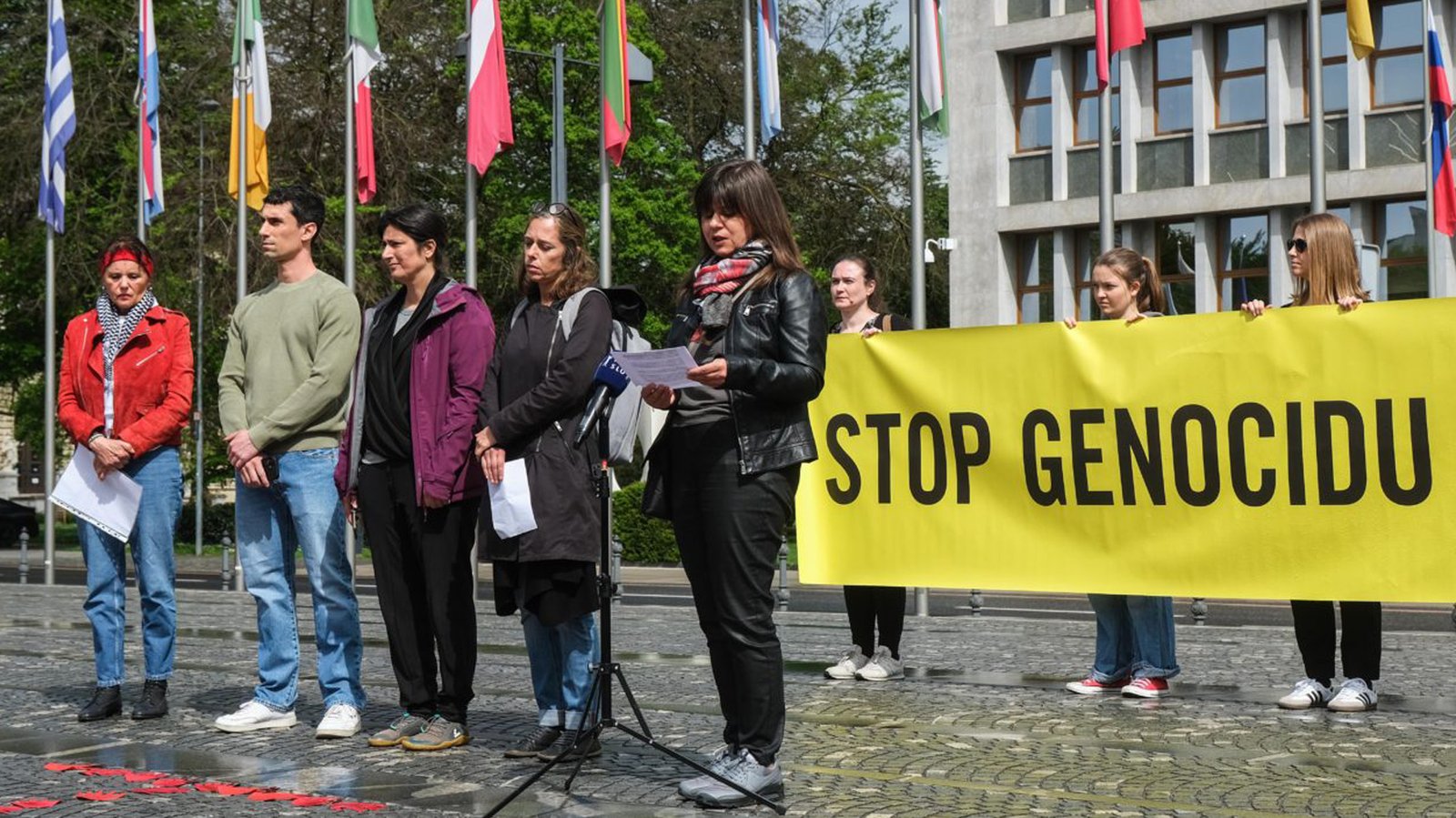 Fotografija predstavnic in predstavnika organizacij, ki so na novinarski konferenci pred DZ predstavile peticijo, v ozadju rumen transparent z napisom Stop genocidu