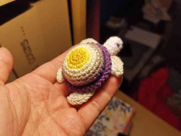 Une main qui tient une tortue crocheté en amigurumi. La carapace de la tortue est composée des couleurs du drapeau non-binaire (jaune, blanc, violet, noir).