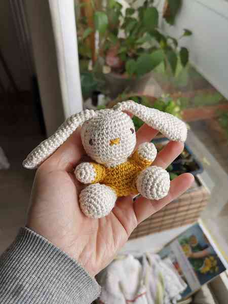 Une main qui tient une mini-peluche lapin, crocheté en amigurumi. Le lapin est blanc avec un vêtement jaune éclatant. On aperçoit des plantes en arrière-plan.