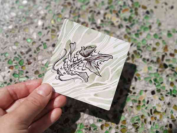 Une main qui tient une illustration bichrome à l'encre calligraphique au-dessus d'une table en mosaïque. L'illustration représente une créature de l'eau proche d'un poisson-chat aux traits très stylisés.