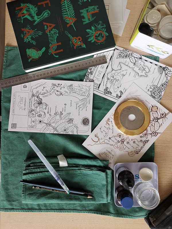 Vue du dessus d'un plan de travail avec plusieurs cartes à l'encre calligraphique, un livre à la couverture verte, un trace cercle et du matériel de dessin.