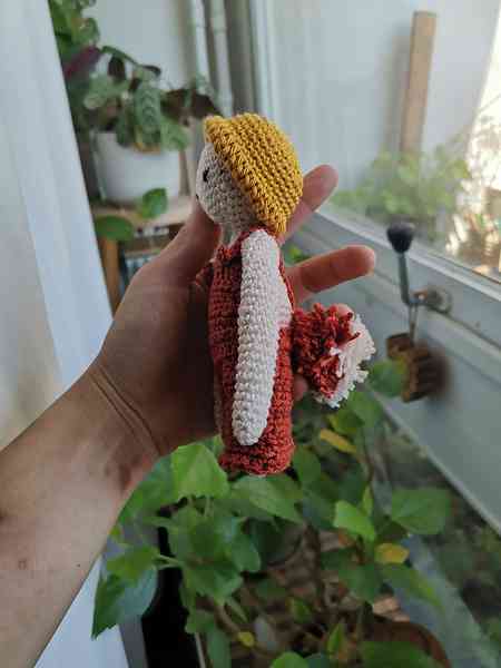 Une main qui tient une mini-peluche, crocheté en amigurumi. C'est un petit personnage tout blanc avec une queue de renard et une salopette orange. On aperçoit des plantes en arrière-plan.