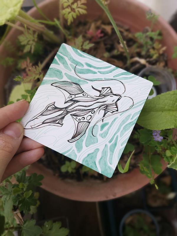 Une main qui tient une illustration bichrome à l'encre calligraphique au-dessus de quelques plantes. L'illustration représente une créature de l'eau proche d'un dragon aux motifs stylisés.