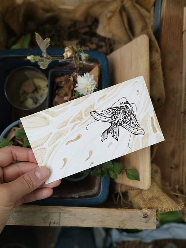 Une main qui tient une illustration bichrome à l'encre calligraphique au-dessus de quelques plantes. L'illustration représente une créature de l'eau proche d'une raie avec une peau aux motifs stylisés.