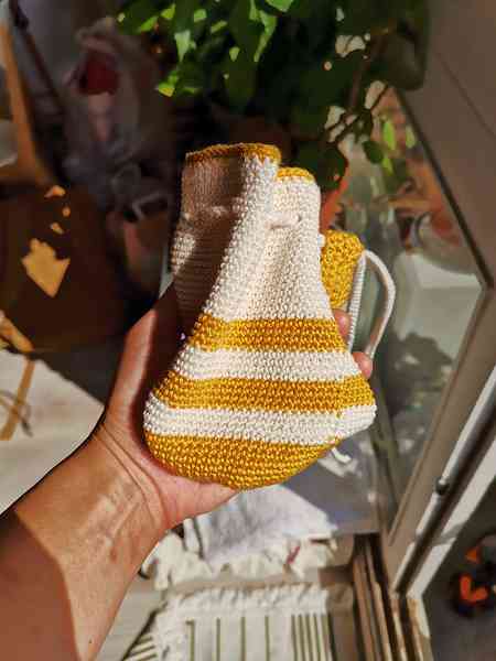 Une main qui tient un sac en crochet. Le sac présente des rayures jaunes et blanches. On aperçoit des plantes en arrière-plan.