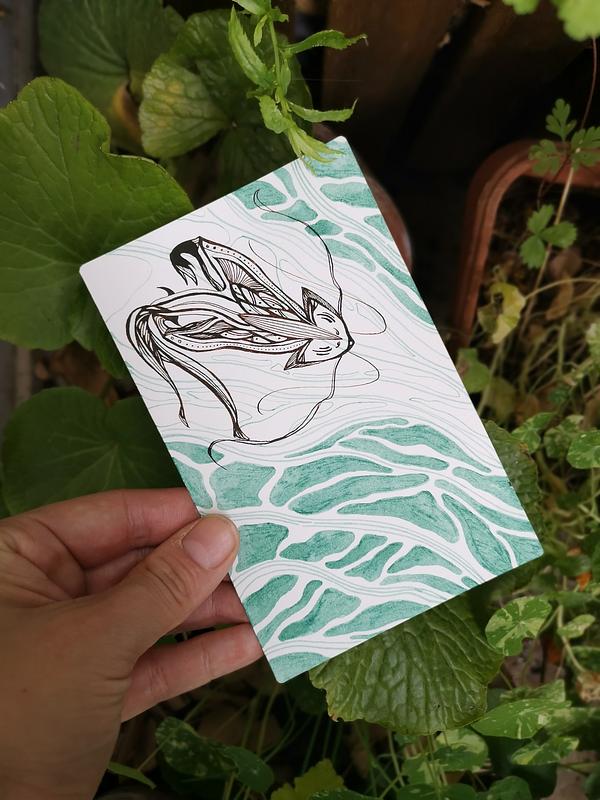 Une main qui tient une illustration bichrome à l'encre calligraphique au-dessus de quelques plantes. L'illustration représente une créature de l'eau proche d'un renard aux motifs stylisés.