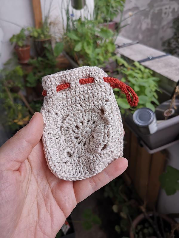 Une main qui tient un mini-pochon beige en crochet avec une corde orange pour le fermer. On aperçoit des plantes en arrière-plan.