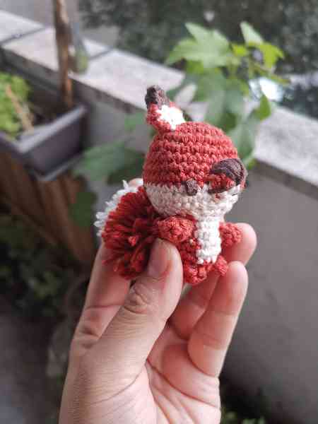 Une main qui tient une mini-peluche renard, crocheté en amigurumi. Le lapin est roux et blanc. On aperçoit des plantes en arrière-plan.