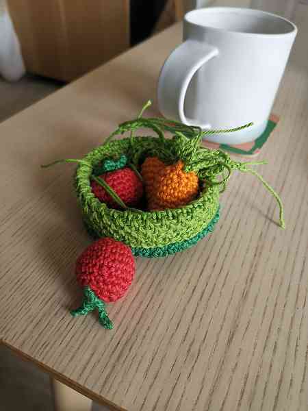 Un petit panier vert en crochet dans lequel sont mis des petites carottes et tomates cerise en amigurumi. Le tout est posé sur une table en bois. On aperçoit une tasse en céramique en arrière-plan.
