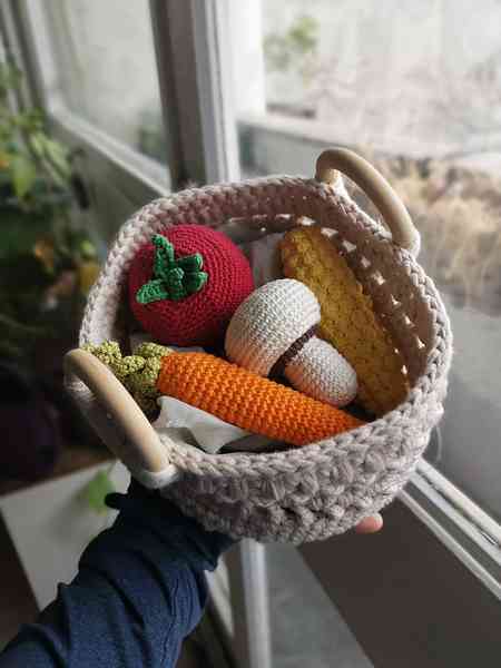 Une main qui tient un panier de couleur blanc cassé avec deux anses en bois. Le panier est crocheté et contient des petits légumes crochetés en amigurumi. On distingue un maïs, une carotte, une tomate et un champignon.
