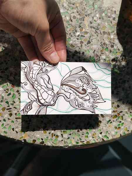 Une main qui tient une illustration bichrome à l'encre calligraphique au-dessus d'une table en mosaïque. L'illustration représente une créature imaginaire proche d'un poisson avec des motifs très stylisés.