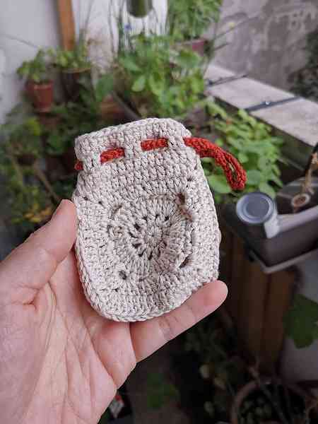 Une main qui tient un mini-pochon beige en crochet avec une corde orange pour le fermer. On aperçoit des plantes en arrière-plan.