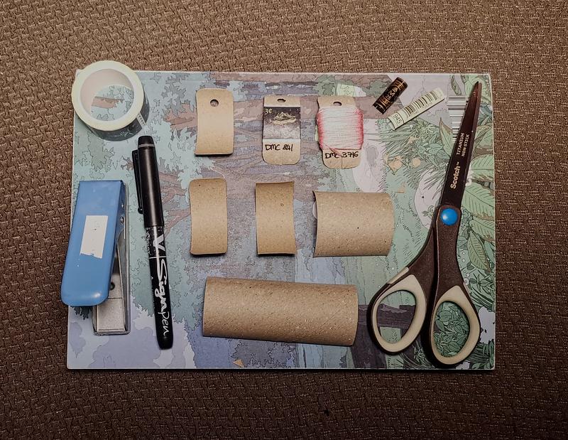 Vue du dessus de morceaux de carton de papier toilette posés les uns à côté des autres montrant les étapes pour fabriquer un petit support pour fil à broder. On aperçoit également une paire de ciseaux, une petite perforeuse, un feutre noir et du masking tape.