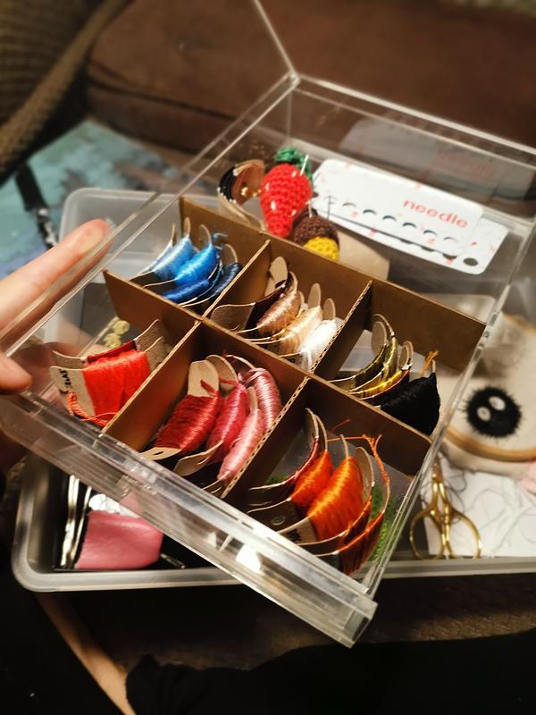 Une main qui tient une boîte transparente dans laquelle sont rangés des fils à broder dans des petites cases en carton. On distingue également une petite fraise et un gland, les deux en crochet sur lesquels sont plantés des aiguilles. 