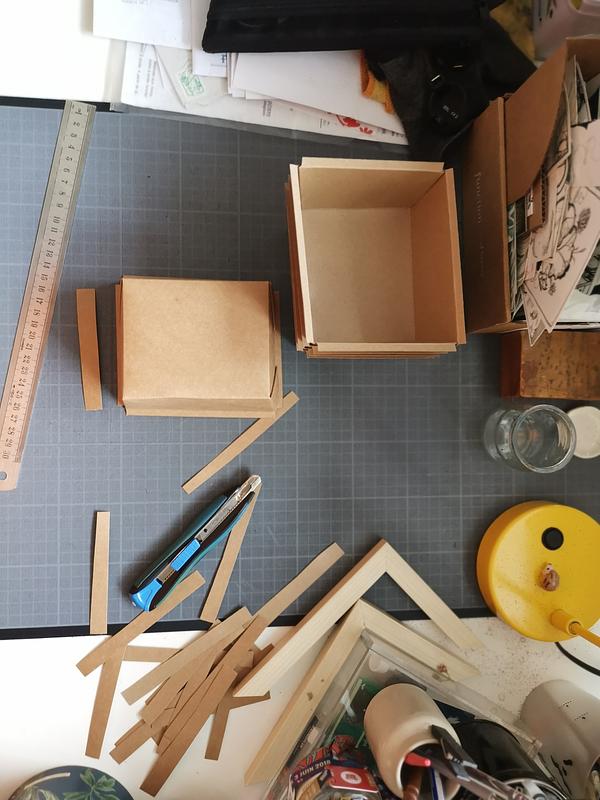 Un bureau vu du dessus avec une planche à couper en lino. On voit des boîtes en carton étalées dessus, ainsi que de nombreuses chutes de carton autour.