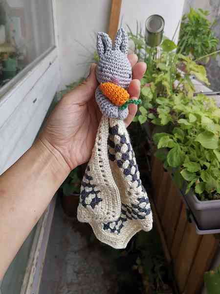 Une main qui tient un doudou lapin. La tête est en crochet version amigurumi et les bras entoure une mini-carotte et une morceau de tissu crocheté en dentelle. On aperçoit des plantes en arrière-plan.