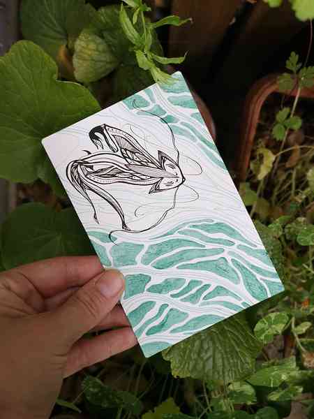 Une main qui tient une illustration bichrome à l'encre calligraphique au-dessus de quelques plantes. L'illustration représente une créature de l'eau proche d'un renard aux motifs stylisés.