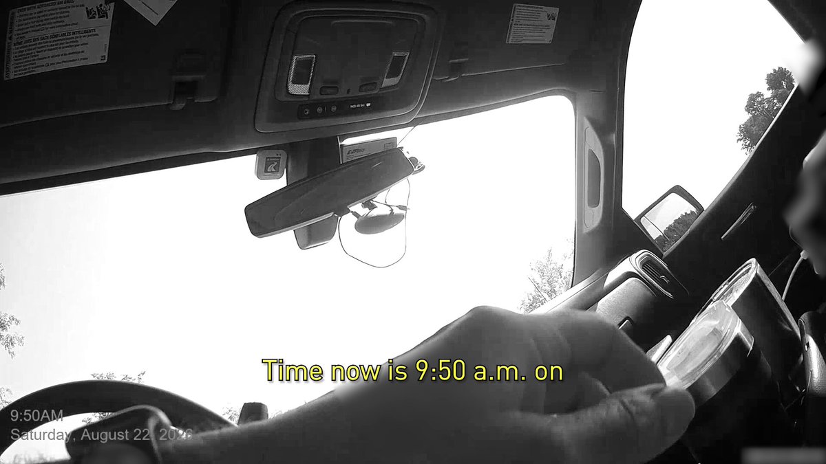 Image en noir et blanc filmée depuis l'intérieur d'une voiture, pare-brise et volant visibles, avec l'horodatage « 9:50AM Saturday, August 22, 2026 » incrusté en bas de l'écran.